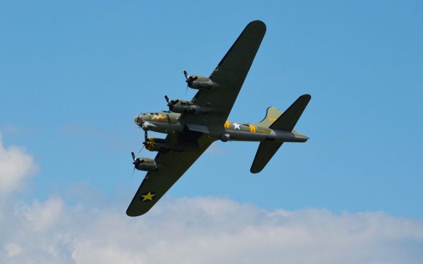 A classic B-17 bomber airplane flying through a clear blue sky with clouds visible.
